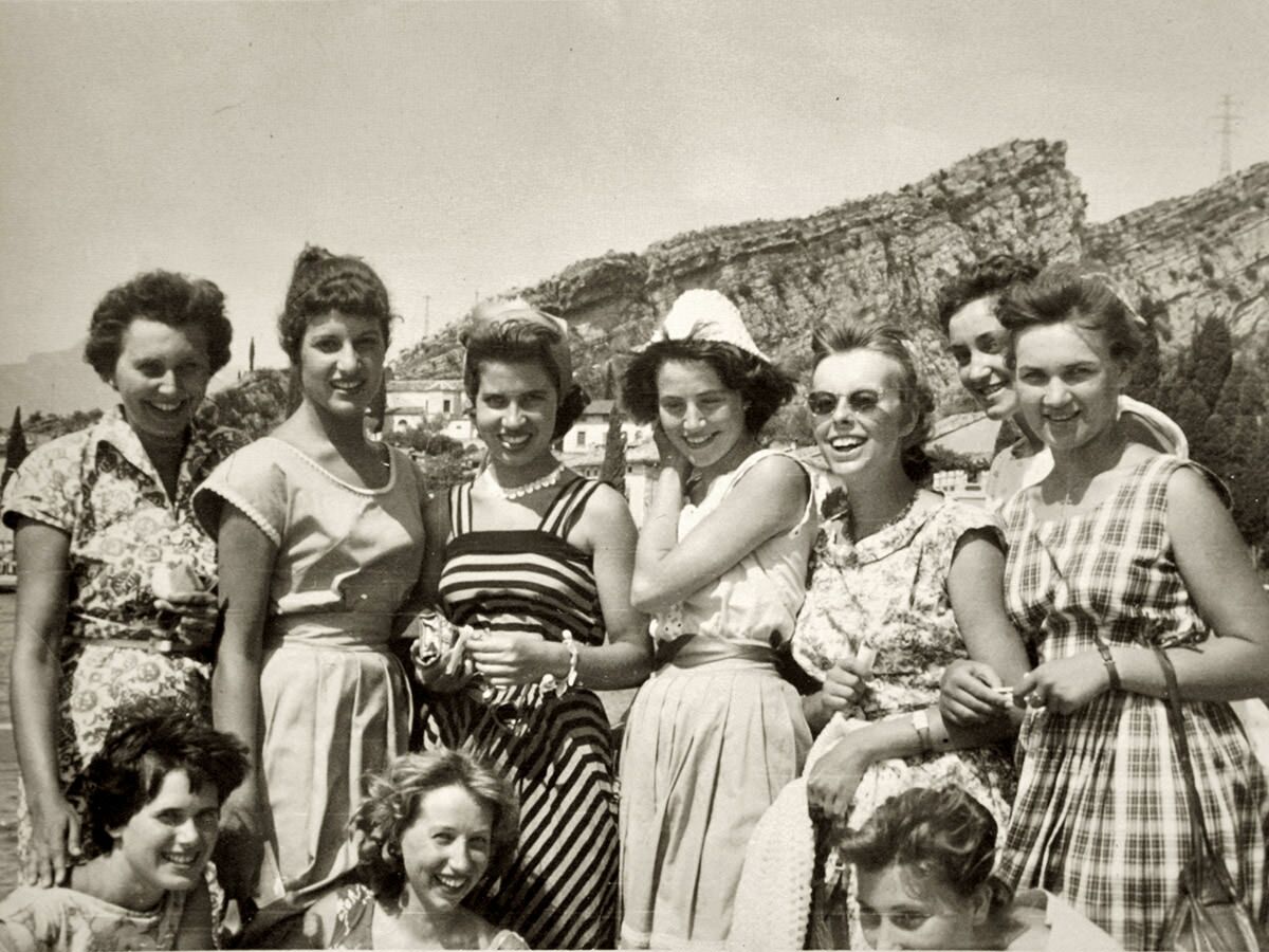 Schwarz-Weiß-Foto einer großen Gruppe lächelnder junger Frauen in Sommerkleidung der 1950er Jahre vor Bergen.