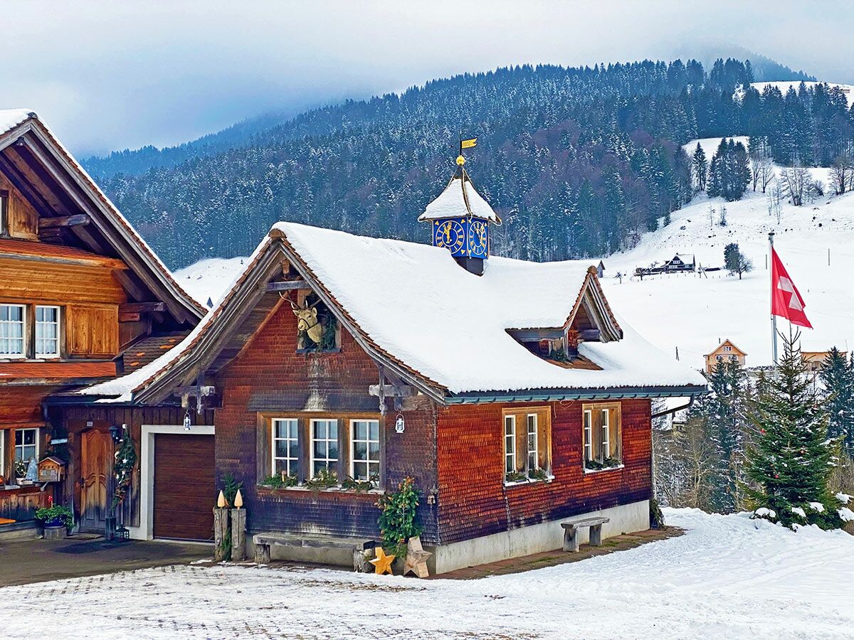 Schneebedeckte traditionelle Schweizer Holzhäuser mit Glockenturm und Hirschkopf-Dekoration in malerischer Alpenlandschaft mit Tannenbäumen und Schweizer Flagge.