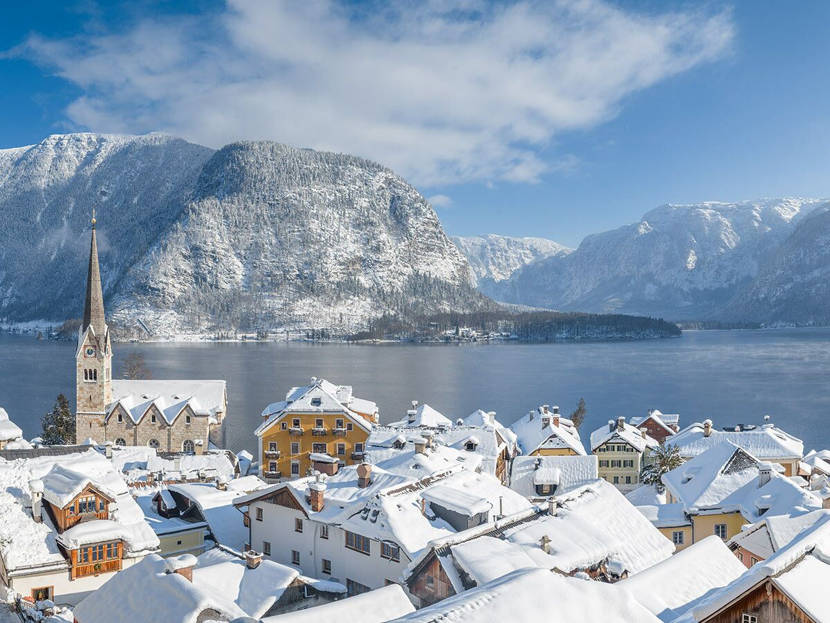 Winterlandschaft von Hallstatt in Österreich mit verschneiten Häusern, Kirche, Hallstätter See und schneebedeckten Bergen unter blauem Himmel.