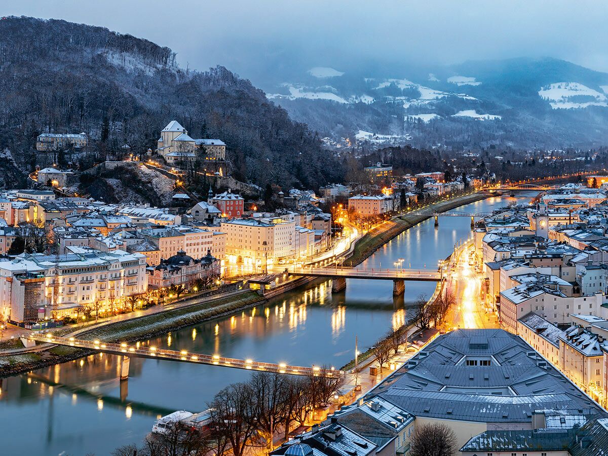 Panoramablick auf das winterliche Salzburg bei Abenddämmerung. Die beleuchtete Salzach schlängelt sich durch die schneebedeckte Stadt.
