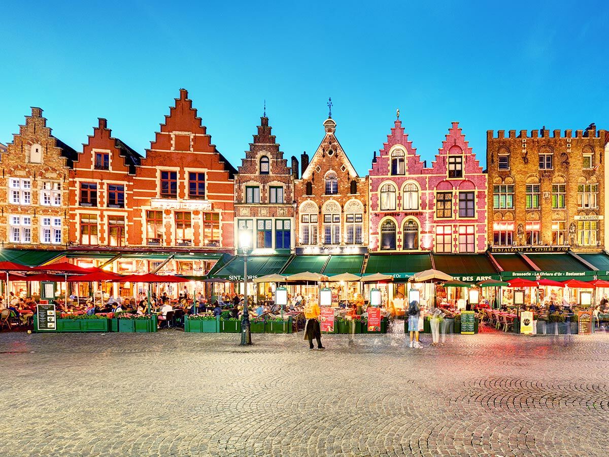 Historischer Marktplatz in Brügge, Belgien, bei Dämmerung mit farbenfrohen Giebelhäusern und belebten Außenrestaurants.