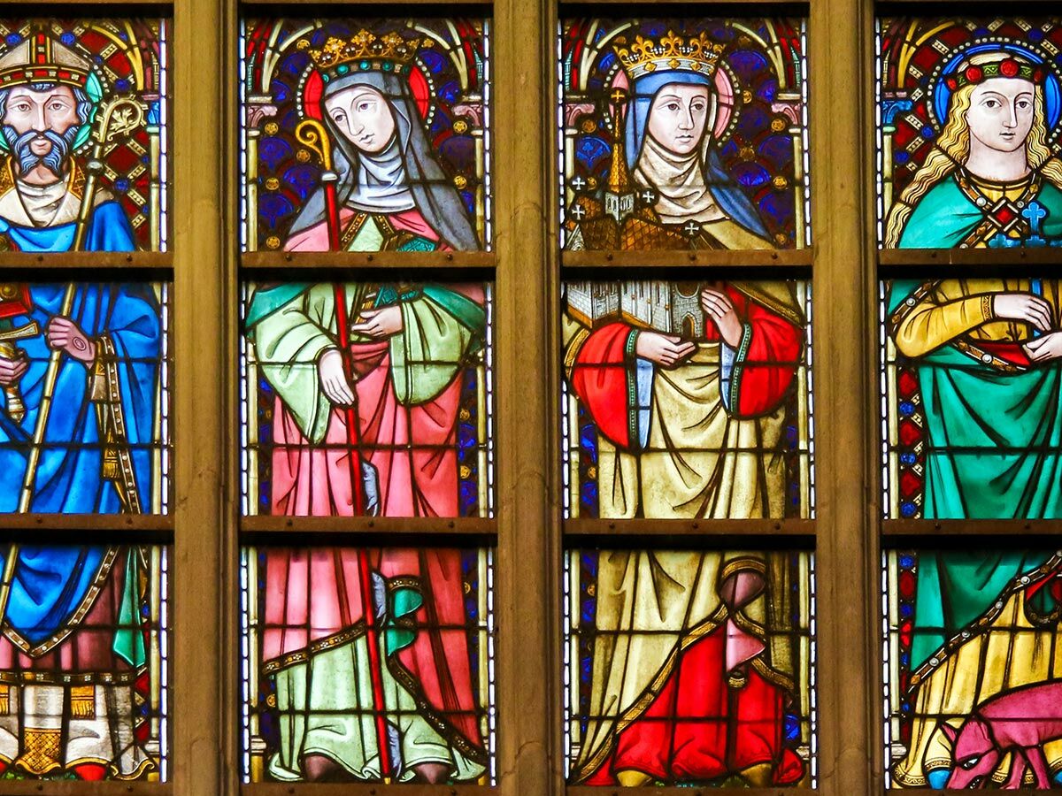 Detailansicht von vier vertikalen Buntglasfenstern in einer Kirche mit Darstellungen von Heiligen, Bischöfen und Nonnen.