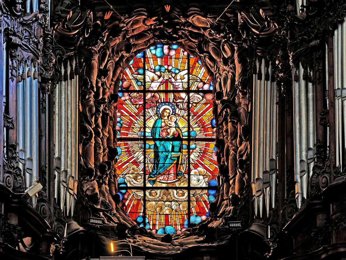 Farbenfrohes Buntglasfenster mit Maria und Jesuskind, gerahmt von kunstvollen Holzschnitzereien und Orgelpfeifen in einer Kirche.