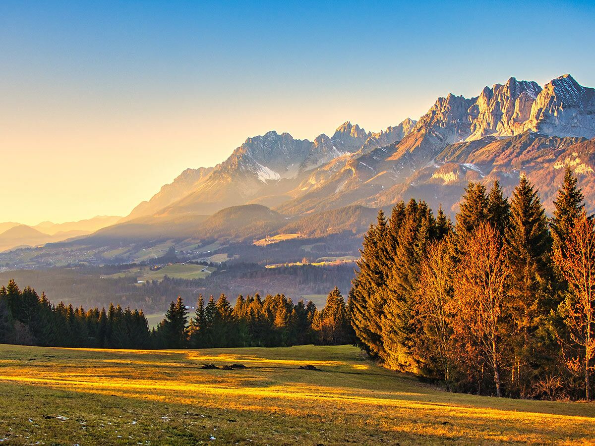Majestätische Alpenlandschaft im goldenen Licht der untergehenden Sonne, mit schneebedeckten Gipfeln, Wäldern und Feldern.