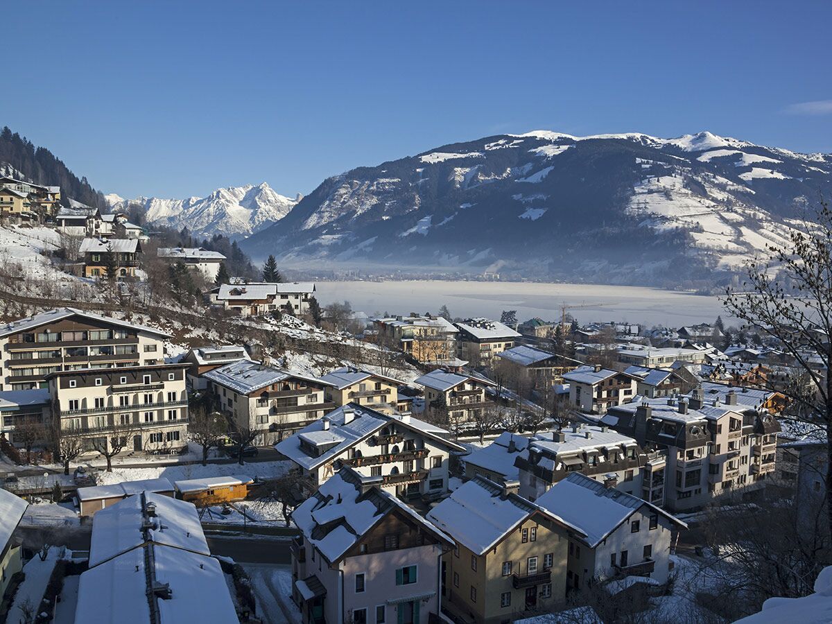 Panoramablick auf ein verschneites Bergdorf mit Häusern am Ufer eines gefrorenen Sees, umgeben von schneebedeckten Alpen.