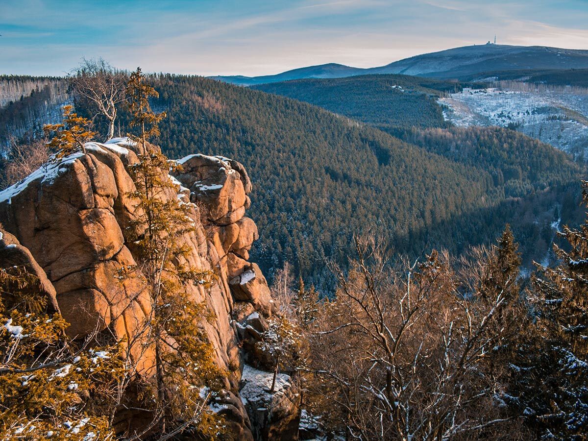 Majestätische Winterlandschaft mit schneebedeckten Felsen im Vordergrund, dichten Wäldern und entfernten Bergen unter blauem Himmel.