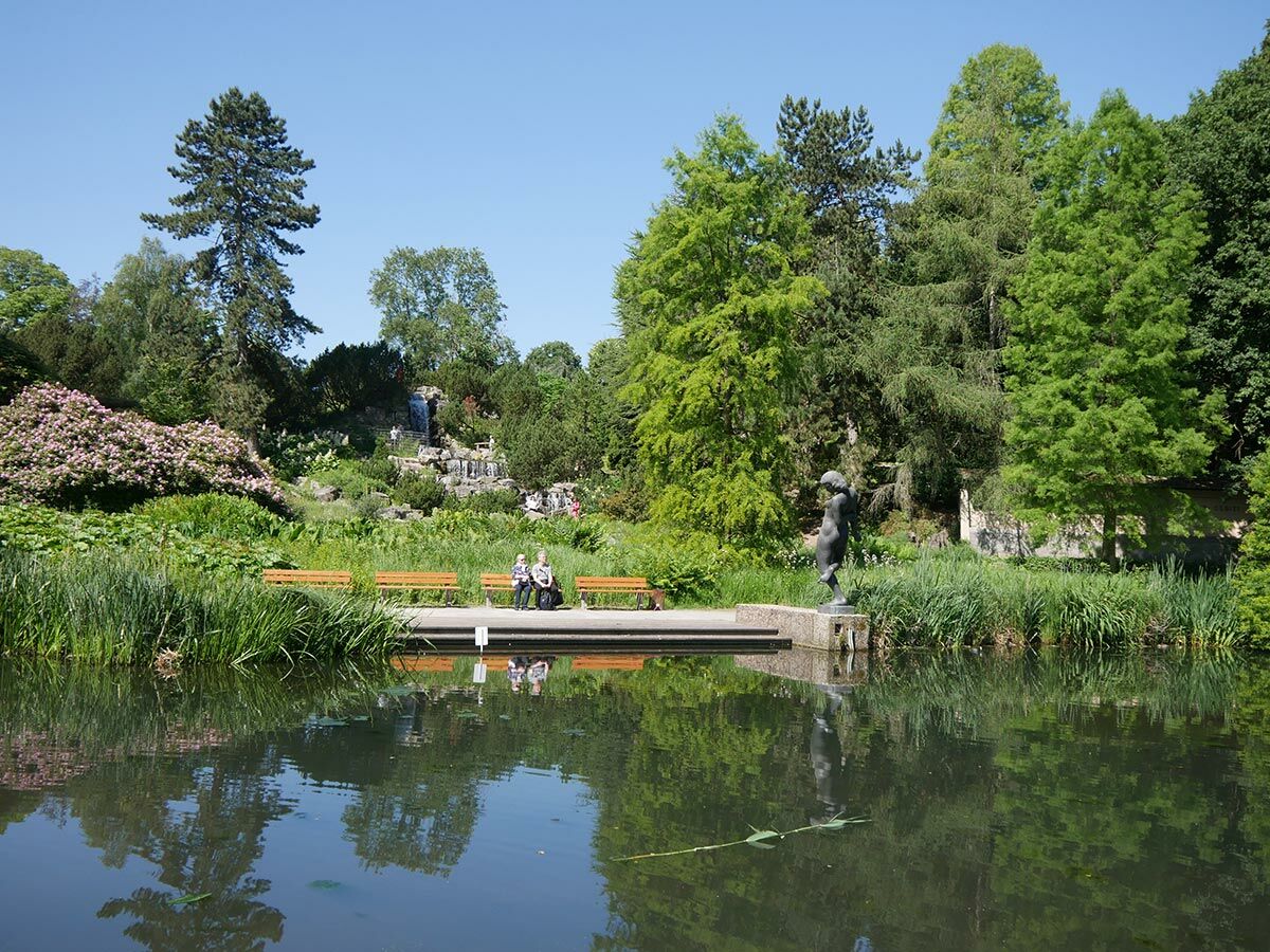 Sonniger Park mit Teich, blühenden Rhododendren, Wasserfall und Nacktstatue am Ufer; Menschen sitzen auf Bänken.