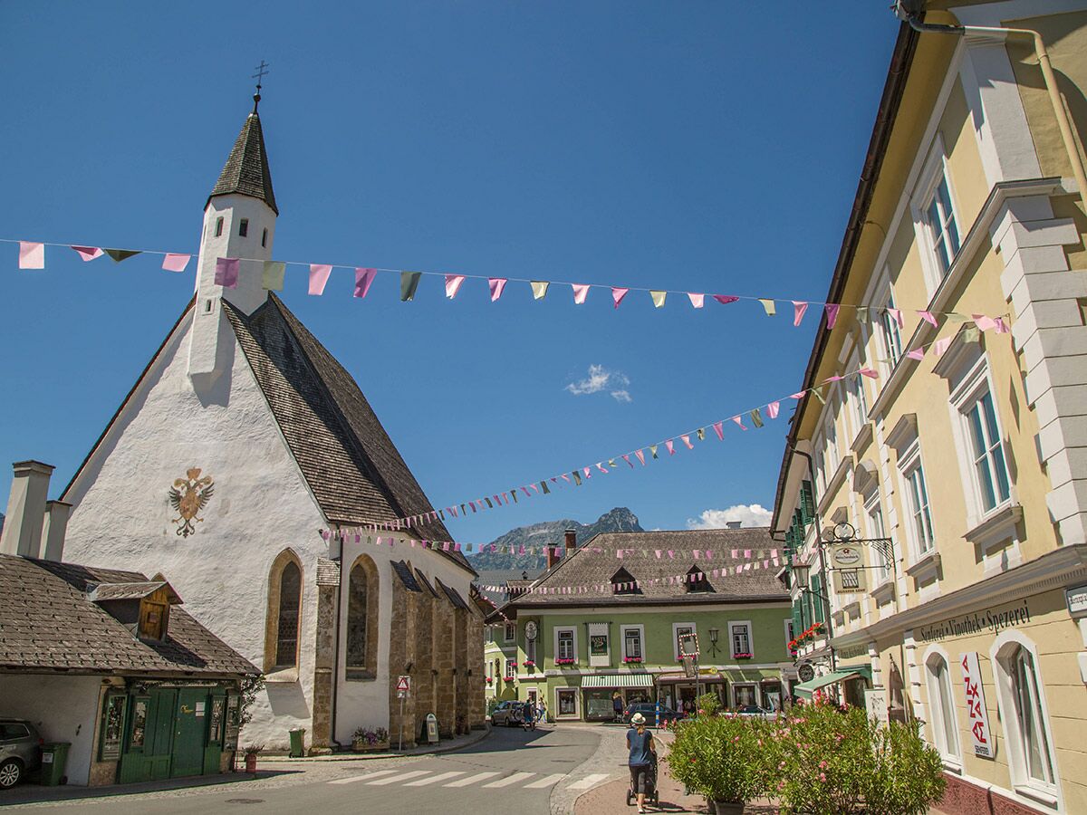 Malerische Dorfstraße mit weißer Kirche, gelben Häusern, bunten Festtagsbannern und Bergen im Hintergrund unter blauem Himmel.