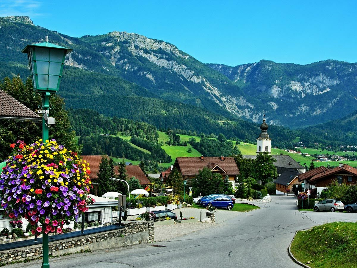 Idyllisches alpines Dorf in Österreich mit bunten Blumen, Kirche, traditionellen Häusern und majestätischen Bergen im Hintergrund.