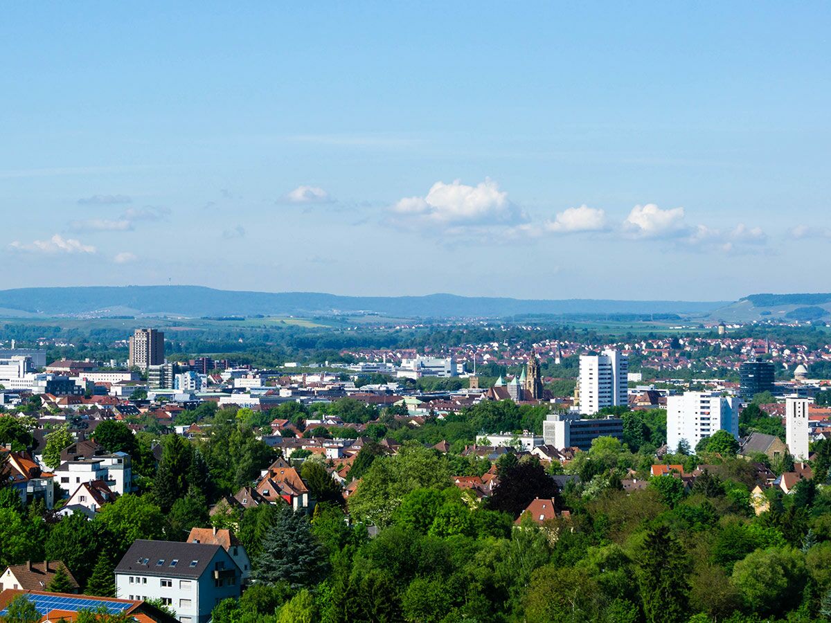 Weitblick über eine Stadt mit viel Grün, Wohnhäusern, Hochhäusern und Kirchen, umrahmt von sanften Hügeln bei blauem Himmel.