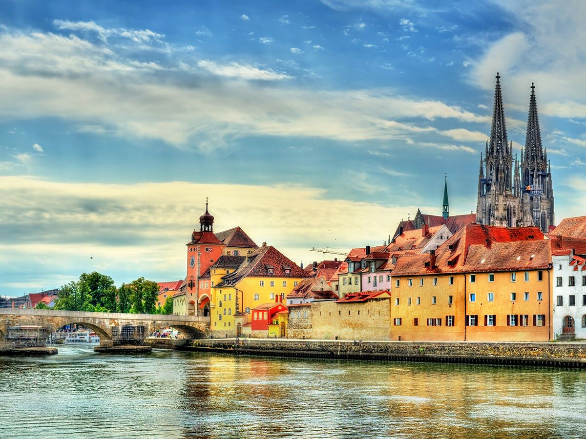 Historische Stadtansicht von Regensburg mit Dom, Steinerner Brücke und bunten Häusern entlang der Donau unter bewölktem Himmel.