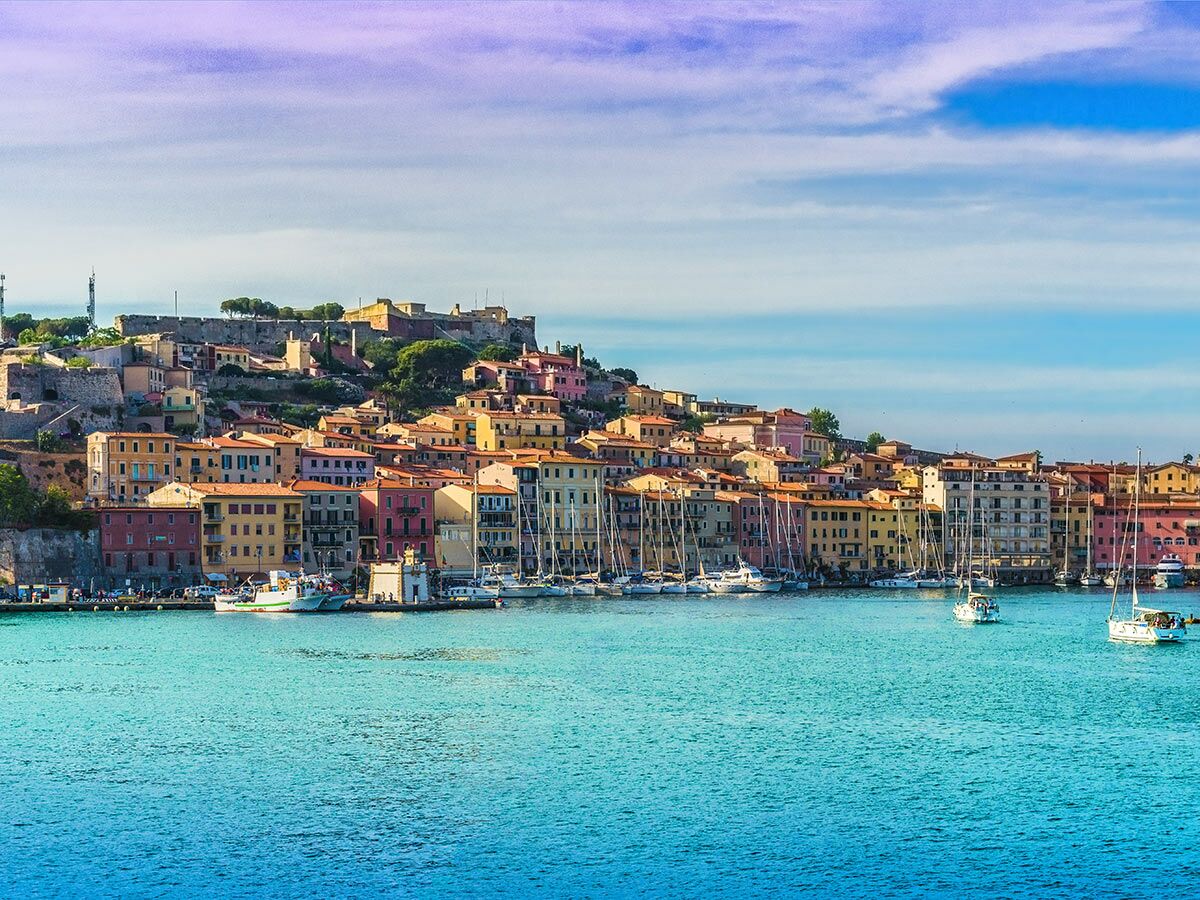 Blick auf Portoferraio, Elba: bunte Häuser am Hang, Segelboote im türkisen Hafenbecken, Festung unter blauem Himmel.