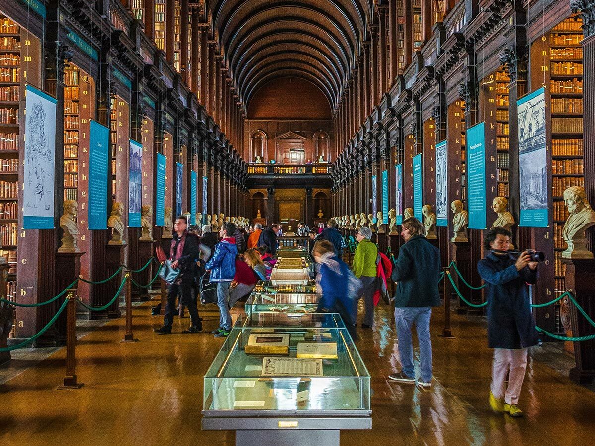 Besucher im majestätischen Langen Saal der Trinity College Bibliothek Dublin mit hohen Bücherregalen, Büsten, Exponaten.