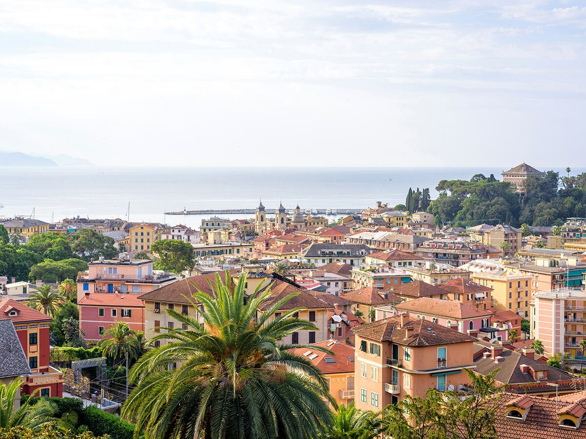 Panoramablick auf eine sonnige italienische Küstenstadt mit bunten Häusern, Palmen, einem Hafen und dem glitzernden Mittelmeer.