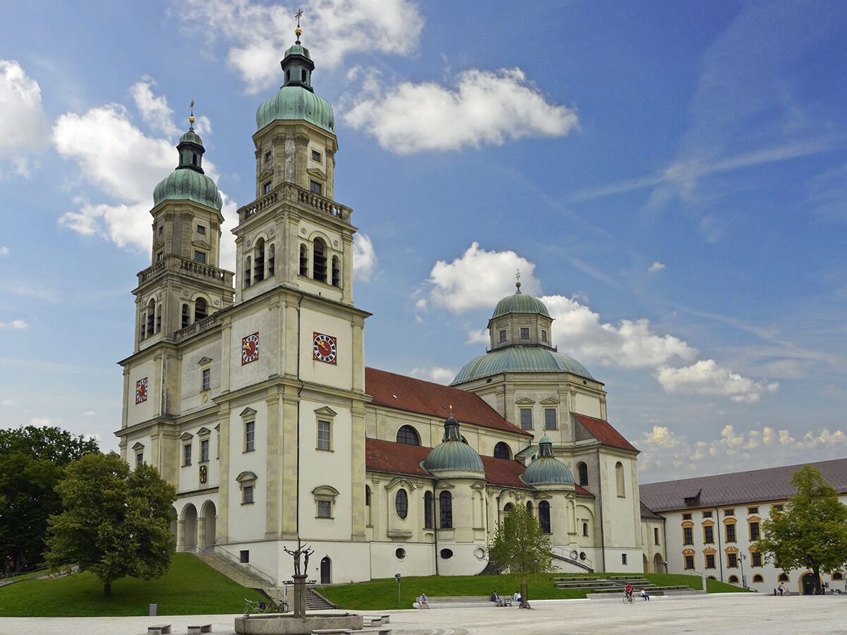 Große barocke Abteikirche Ottobeuren mit zwei Türmen, Kuppeln und rotem Dach unter blauem Himmel auf einem sonnigen Platz.