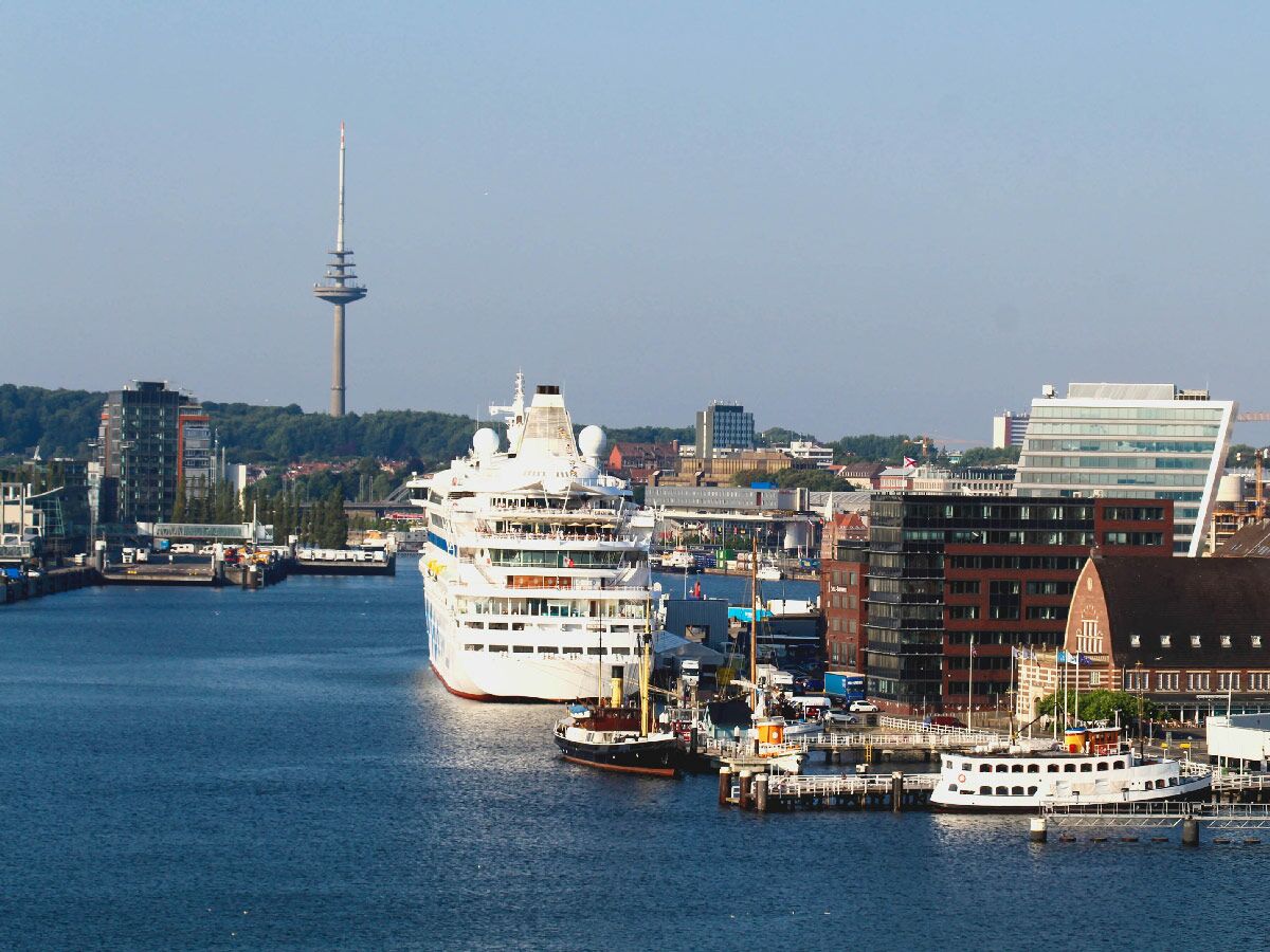 Ein großes AIDA-Kreuzfahrtschiff im Kieler Hafen, umgeben von Stadtgebäuden, kleinen Booten und dem markanten Fernsehturm.