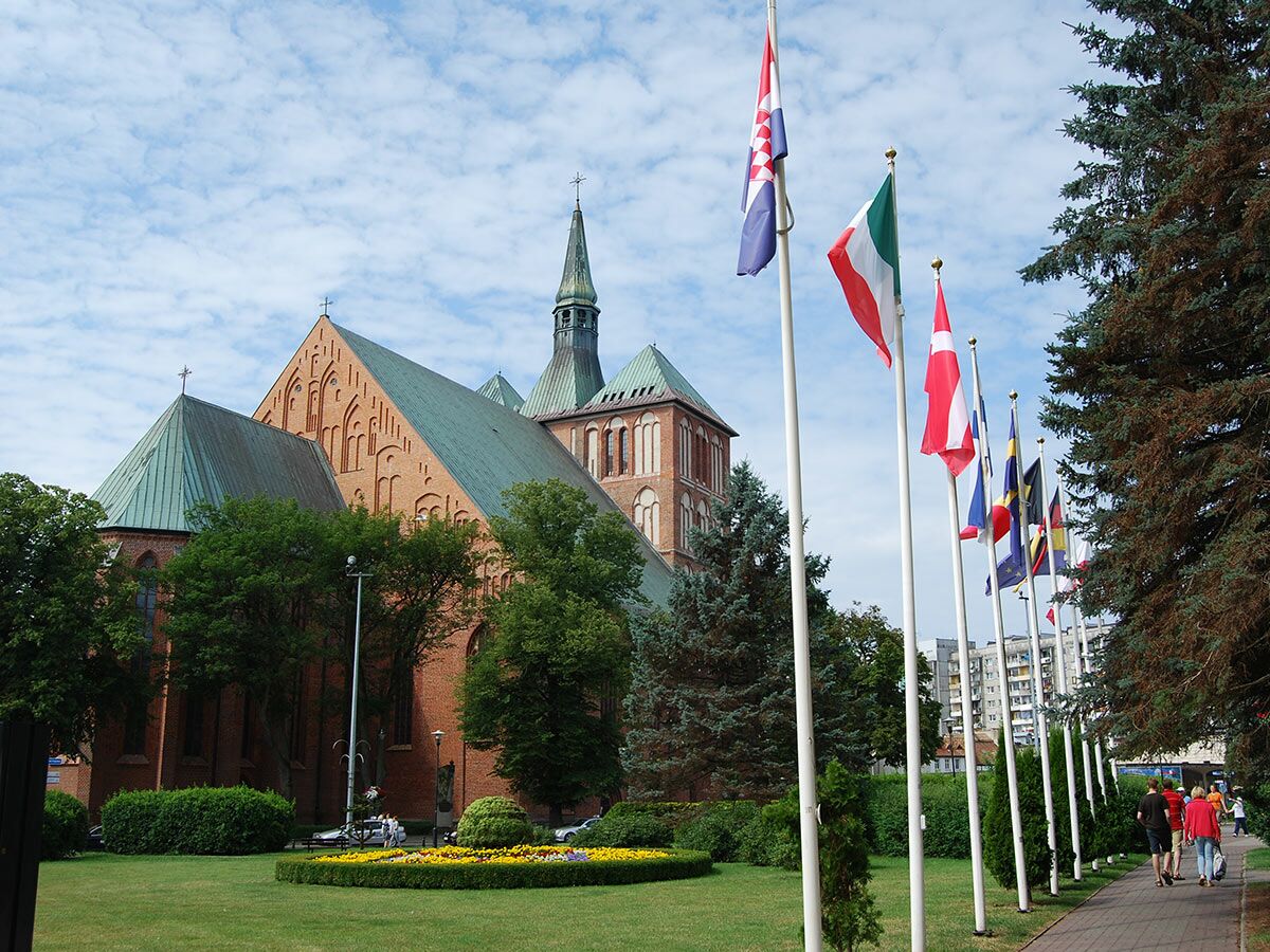 Historische gotische Backsteinkirche mit grünem Dach und Turm, umgeben von einem Park mit Blumen und wehenden europäischen Flaggen unter blauem Himmel.