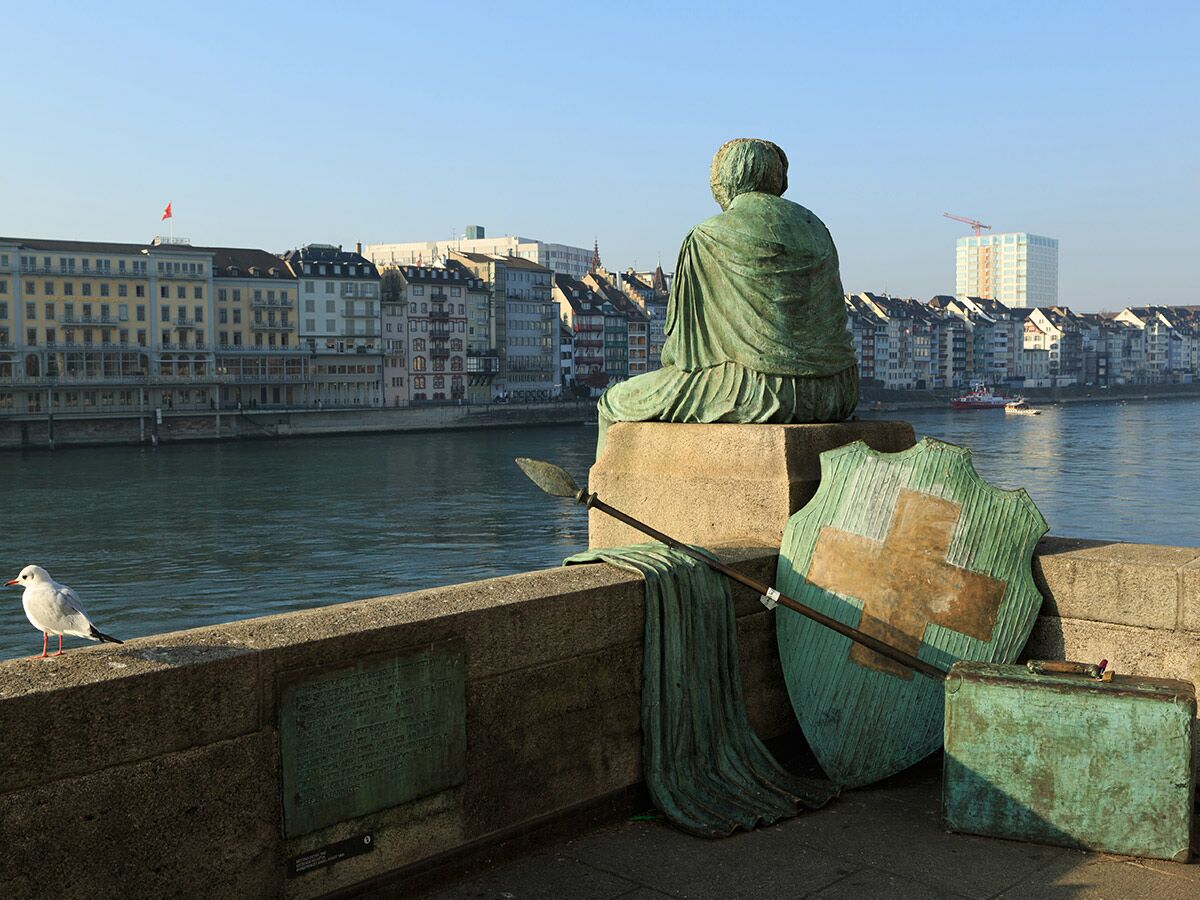 Sitzende Bronzestatue mit Schild, Speer und Koffer am Rhein in Basel, dahinter Fluss und Stadtkulisse.