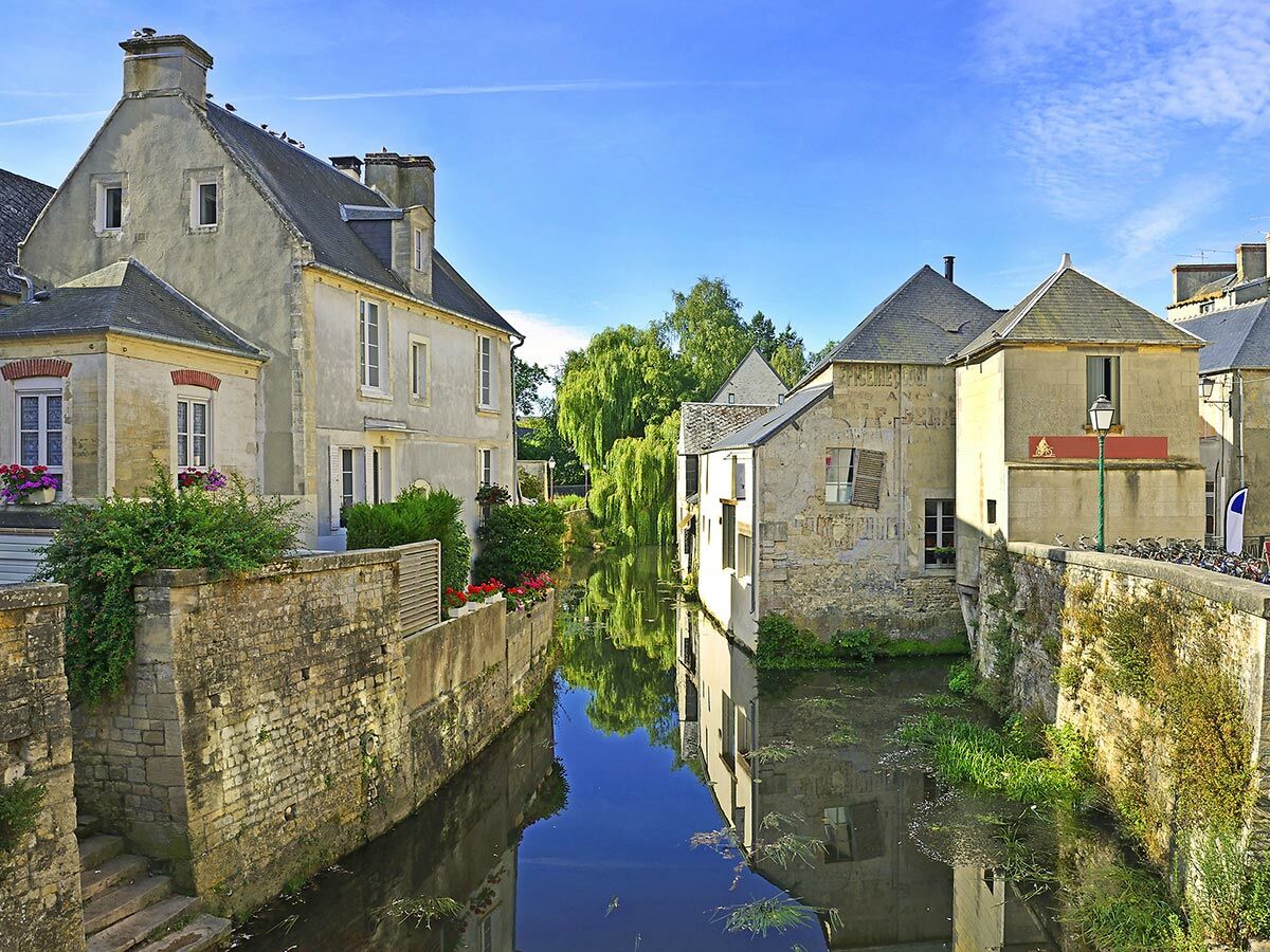 Malerische historische Häuser und Steinmauern spiegeln sich in einem ruhigen Kanal unter blauem Himmel in einer charmanten Altstadt.