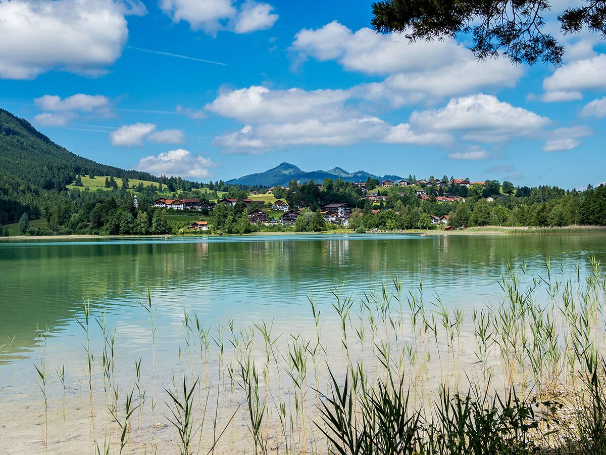 Blick über einen ruhigen See mit Schilf auf ein malerisches Dorf an bewaldeten Hügeln und Bergen unter blauem Himmel.