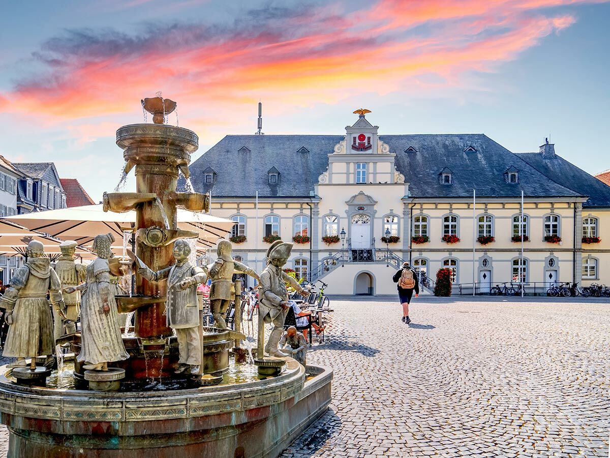 Historisches Rathaus und Brunnen mit Statuen auf Kopfsteinpflasterplatz bei Sonnenuntergang; eine Person geht weg.
