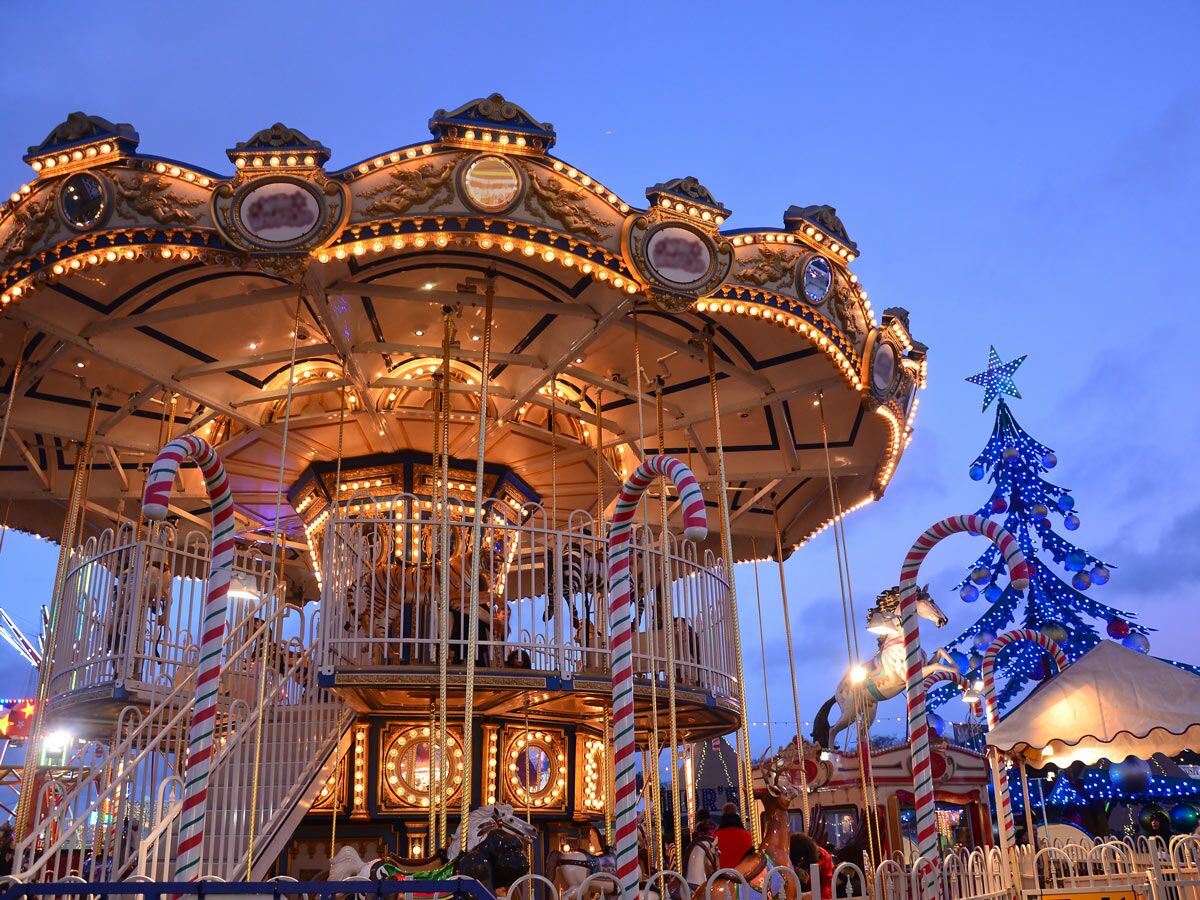 Beleuchtetes Karussell auf einem Weihnachtsmarkt bei Abenddämmerung mit blauem Weihnachtsbaum und festlicher Dekoration.