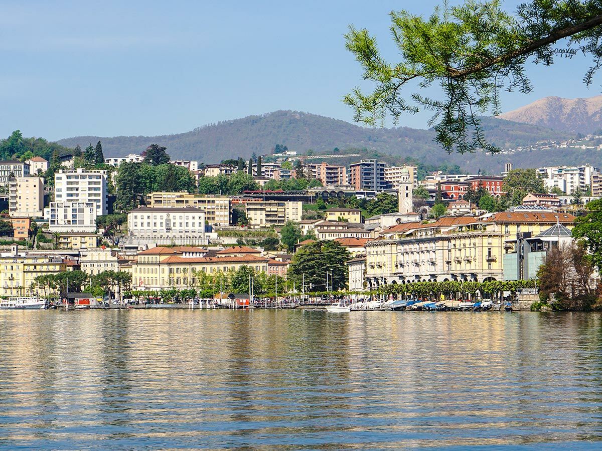 Panoramablick auf Lugano, Schweiz, mit Gebäuden am Ufer des Luganersees und Bergen im Hintergrund unter blauem Himmel.