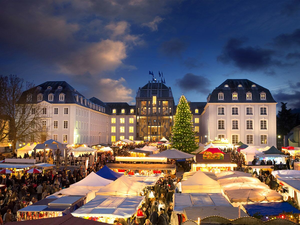 Festlicher Weihnachtsmarkt am Abend mit vielen Besuchern, beleuchteten Ständen, historischen Gebäuden und großem Weihnachtsbaum.