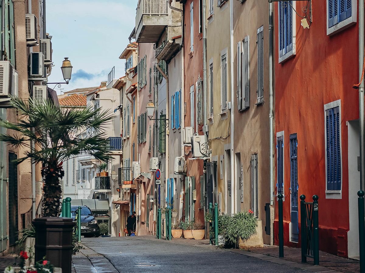 Enge Gasse mit bunten, mediterranen Häusern, blauen Fensterläden und einer Palme am Straßenrand in Südeuropa.