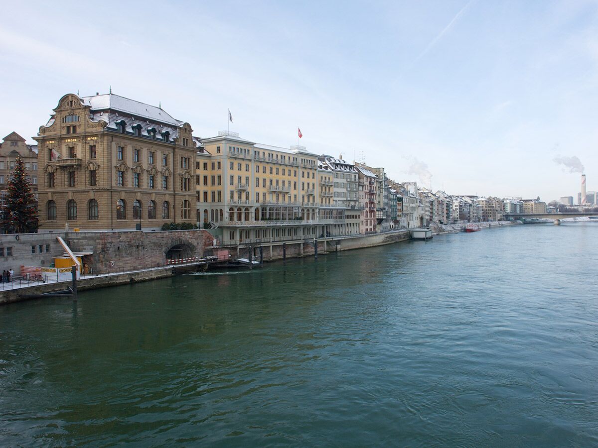 Panoramablick auf Basel am Rhein im Winter mit dem Hotel Les Trois Rois und schneebedeckten Ufergebäuden.