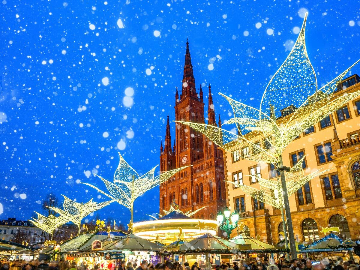 Festlicher Weihnachtsmarkt in Wiesbaden bei Nacht mit fallendem Schnee, beleuchteter Marktkirche, leuchtenden Dekorationen und Glühweinständen.