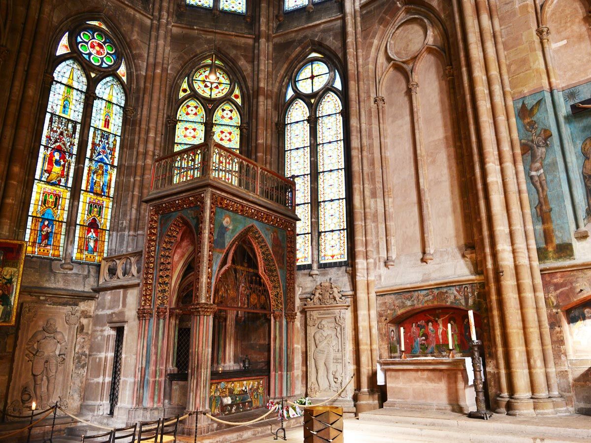 Detailreiches Kircheninneres einer gotischen Kathedrale mit leuchtenden Buntglasfenstern, verzierter Kanzel, Altar und Wandmalereien.