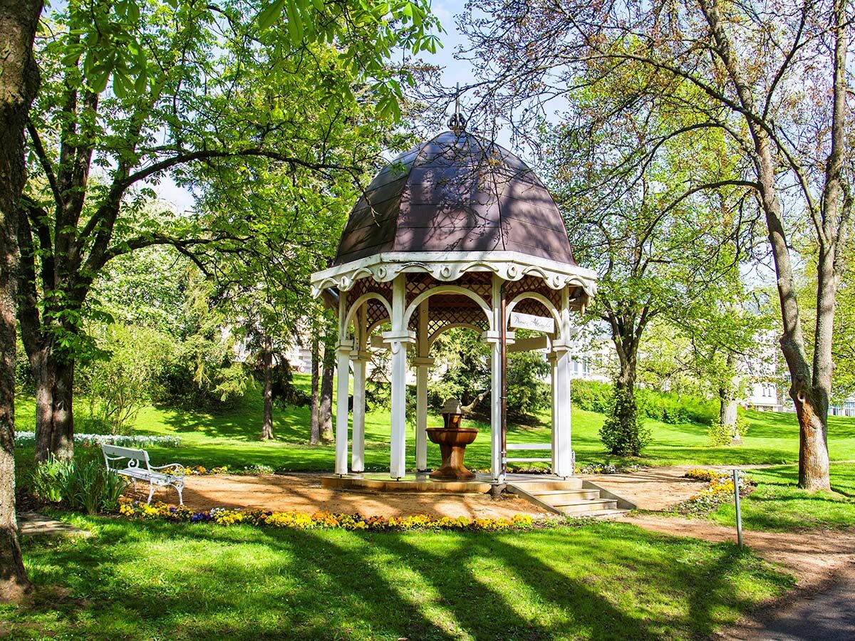 Malerischer Parkpavillon mit Brunnen im grünen Frühlingspark. Zahlreiche Bäume, Rasenflächen, Blumenbeete und eine weiße Parkbank.