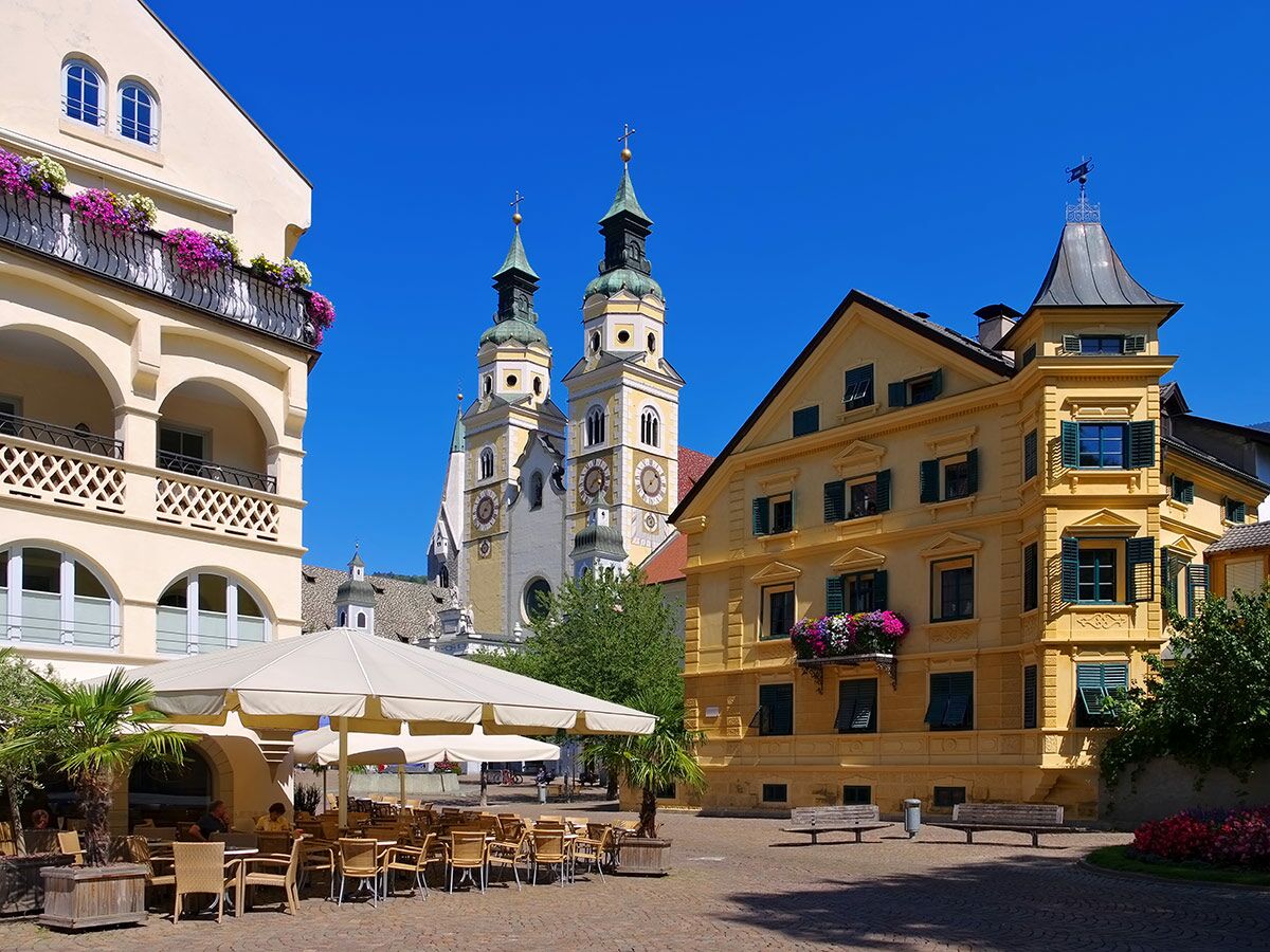 Historischer Platz in Meran, Südtirol, mit alten Gebäuden, blühenden Balkonen, Kirchtürmen und Straßencafé unter blauem Himmel.
