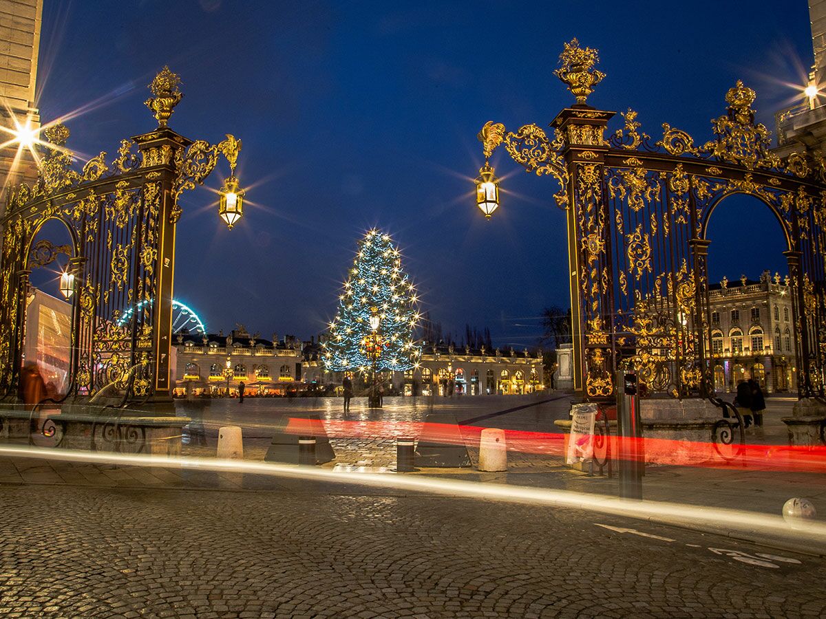 Festlicher Weihnachtsbaum auf dem Place Stanislas in Nancy bei Nacht, umgeben von goldenen Toren und beleuchteten Gebäuden mit Lichtspuren.