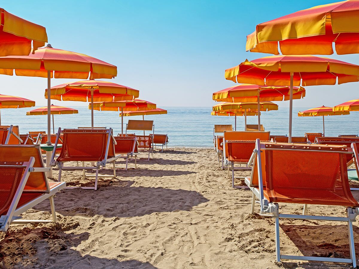 Viele orange-gelbe Sonnenschirme und Liegestühle am sonnigen Sandstrand vor dem blauen Meer.