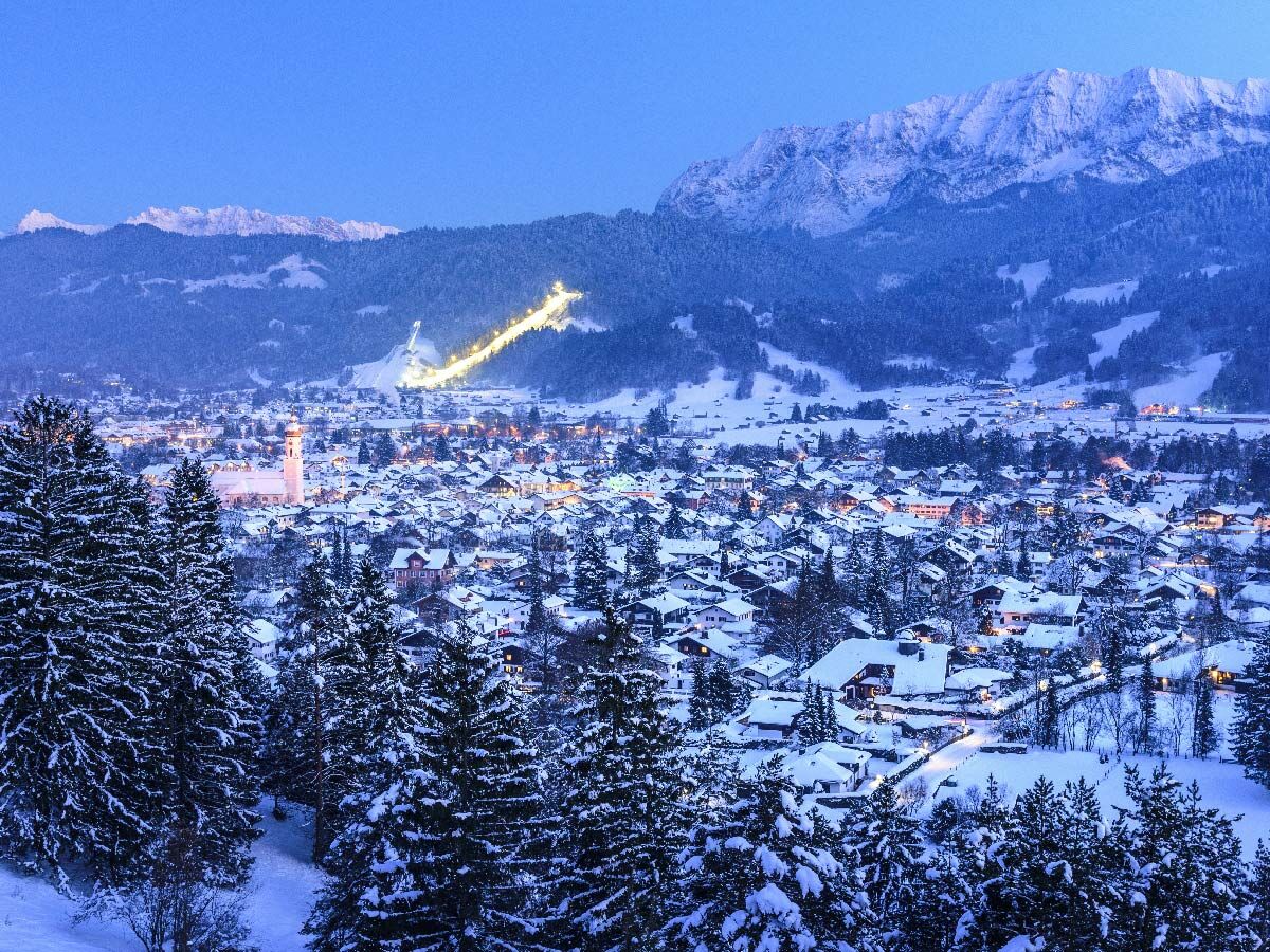 Idyllisches, schneebedecktes Bergdorf mit beleuchteten Häusern, Kirche, Pinien und Alpenkulisse sowie leuchtender Skipiste zur Dämmerung.