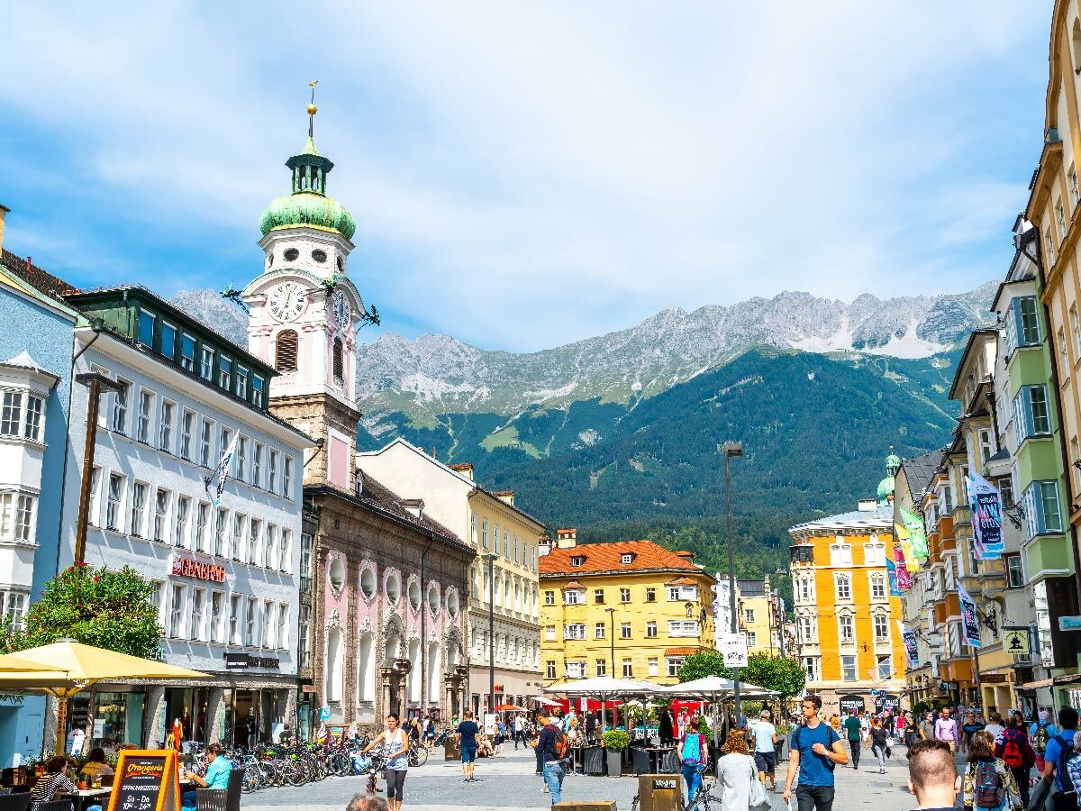 Belebte Altstadt von Innsbruck mit historischen Gebäuden, Kirchturm und der majestätischen Nordkette im Hintergrund bei Sonnenschein.