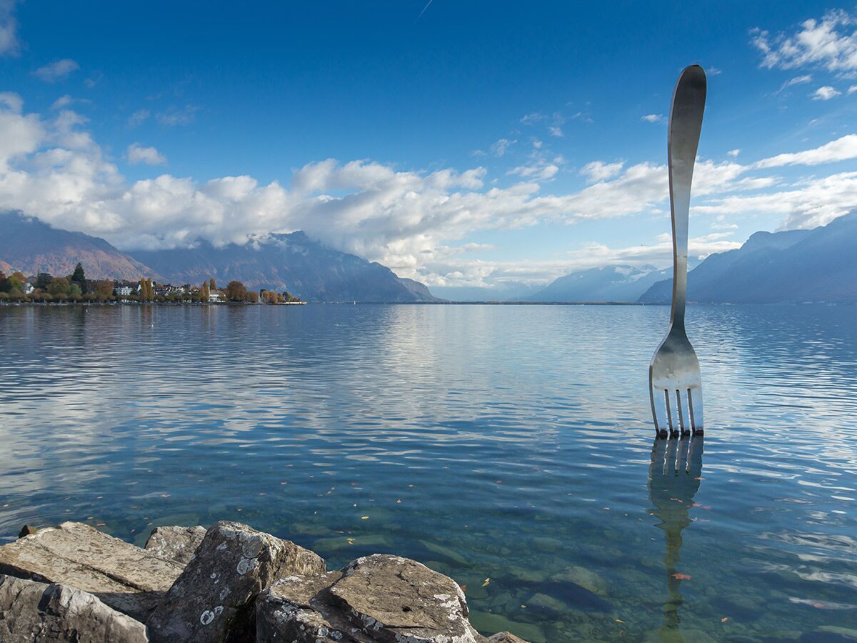 Die berühmte Riesen-Gabel Skulptur im Genfersee bei Vevey, Schweiz, mit Bergen, Himmel und Uferbebauung im Hintergrund.