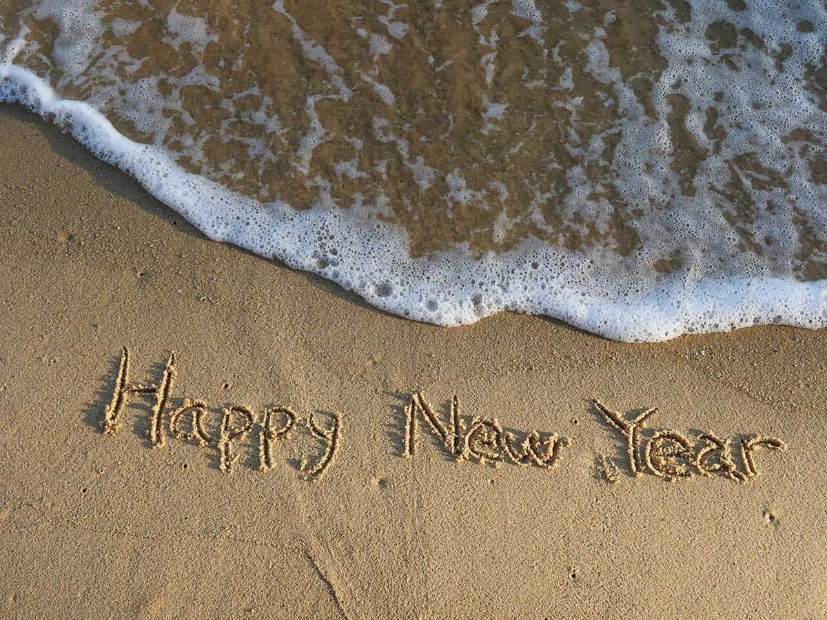 Die Worte "Happy New Year" sind in den goldenen Sand eines Strandes geschrieben, während eine schäumende Meereswelle zurückweicht.