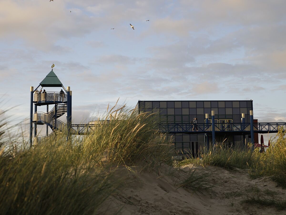 Moderner Aussichtsturm mit Wendeltreppe und Gebäude auf Sanddünen mit Gras und fliegenden Möwen unter bewölktem Himmel.