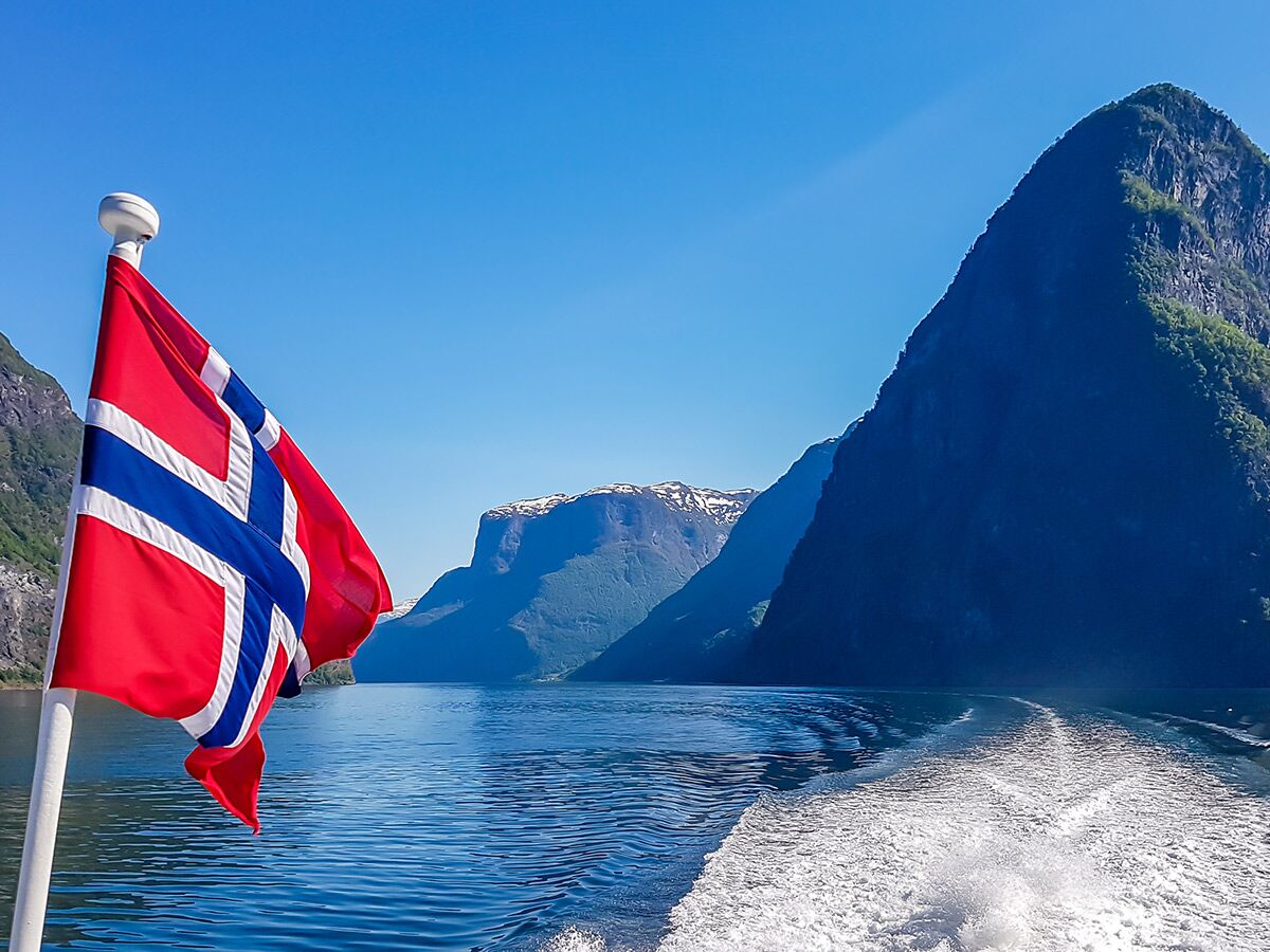 Norwegische Flagge weht an Bord eines Bootes auf einem sonnigen Fjord, umgeben von majestätischen Bergen unter blauem Himmel.