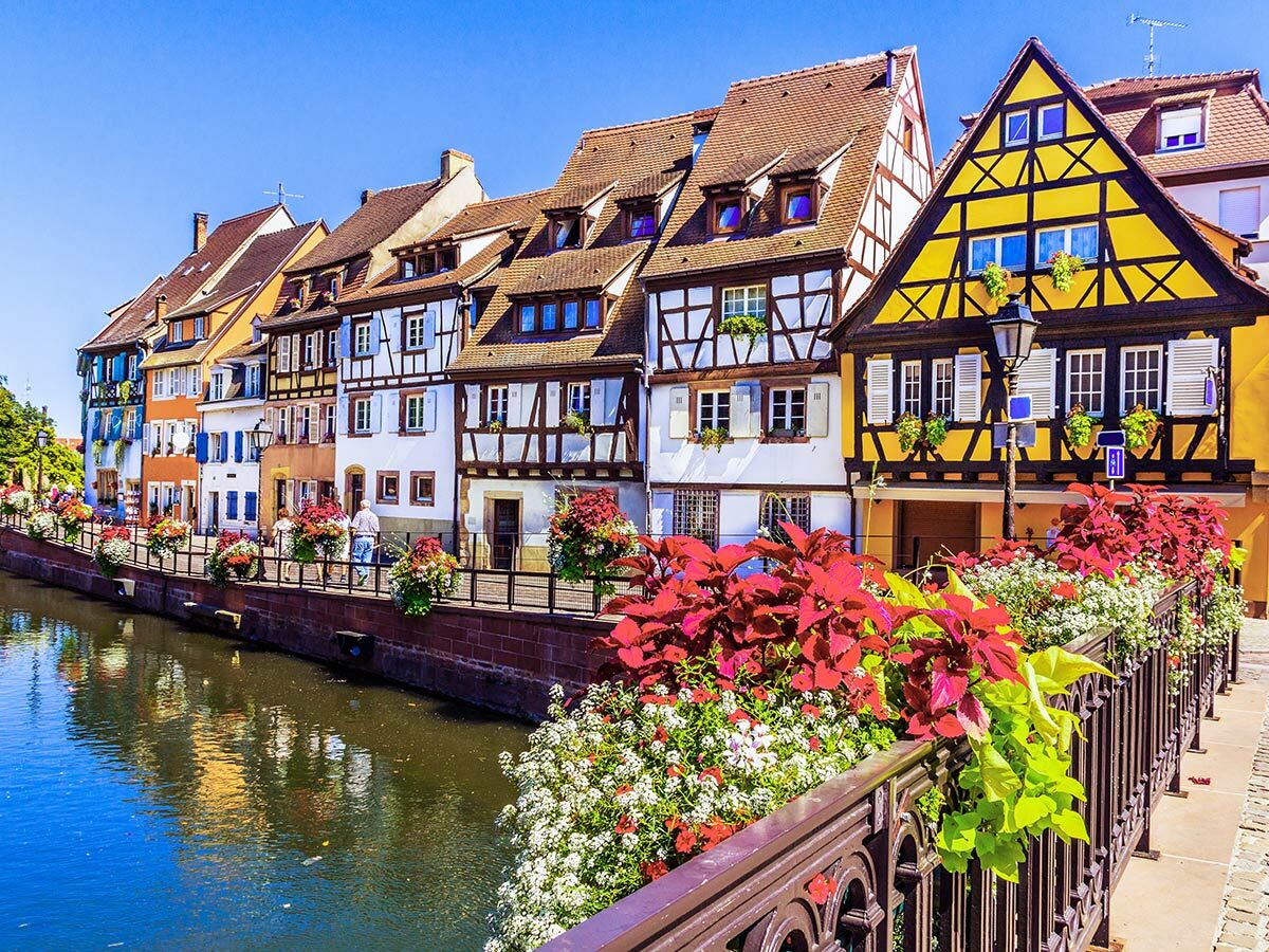 Bunte Fachwerkhäuser am Kanal in Colmar, Elsass, mit Blumen an der Brücke unter blauem Himmel, Reflexionen im Wasser.