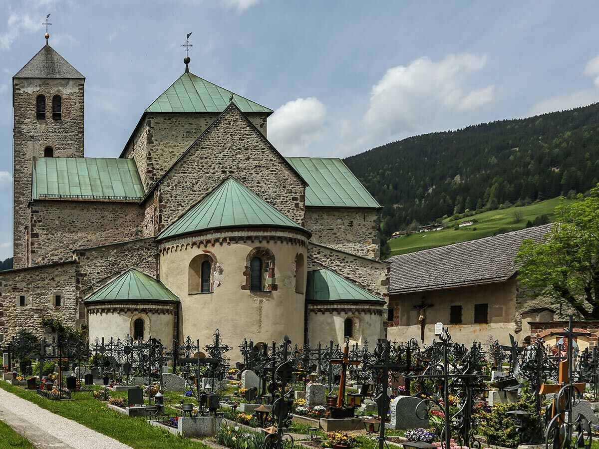 Alte Steinkirche mit grünem Kupferdach und Glockenturm, umgeben von einem Friedhof mit schmiedeeisernen Kreuzen vor bewaldeten Bergen.