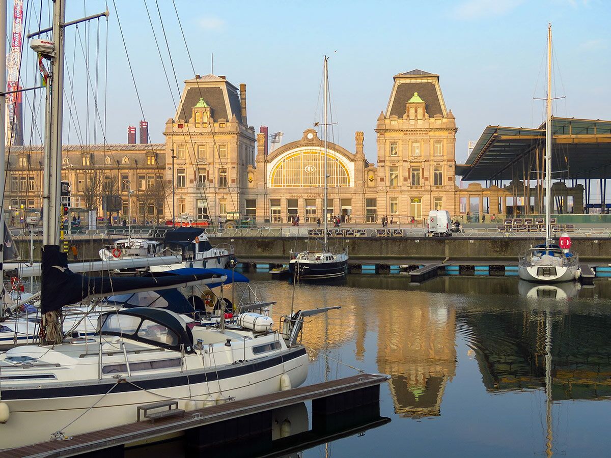 Segelboote im Hafen von Oostende mit der Architektur des historischen Bahnhofsgebäudes und Reflexionen im Wasser.