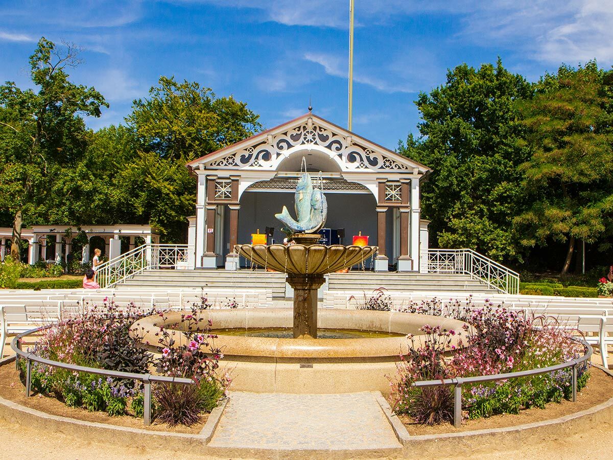 Ein historischer Musikpavillon mit einem Springbrunnen und Blumenbeeten vor weißen Sitzbänken in einem sonnigen Park unter blauem Himmel.