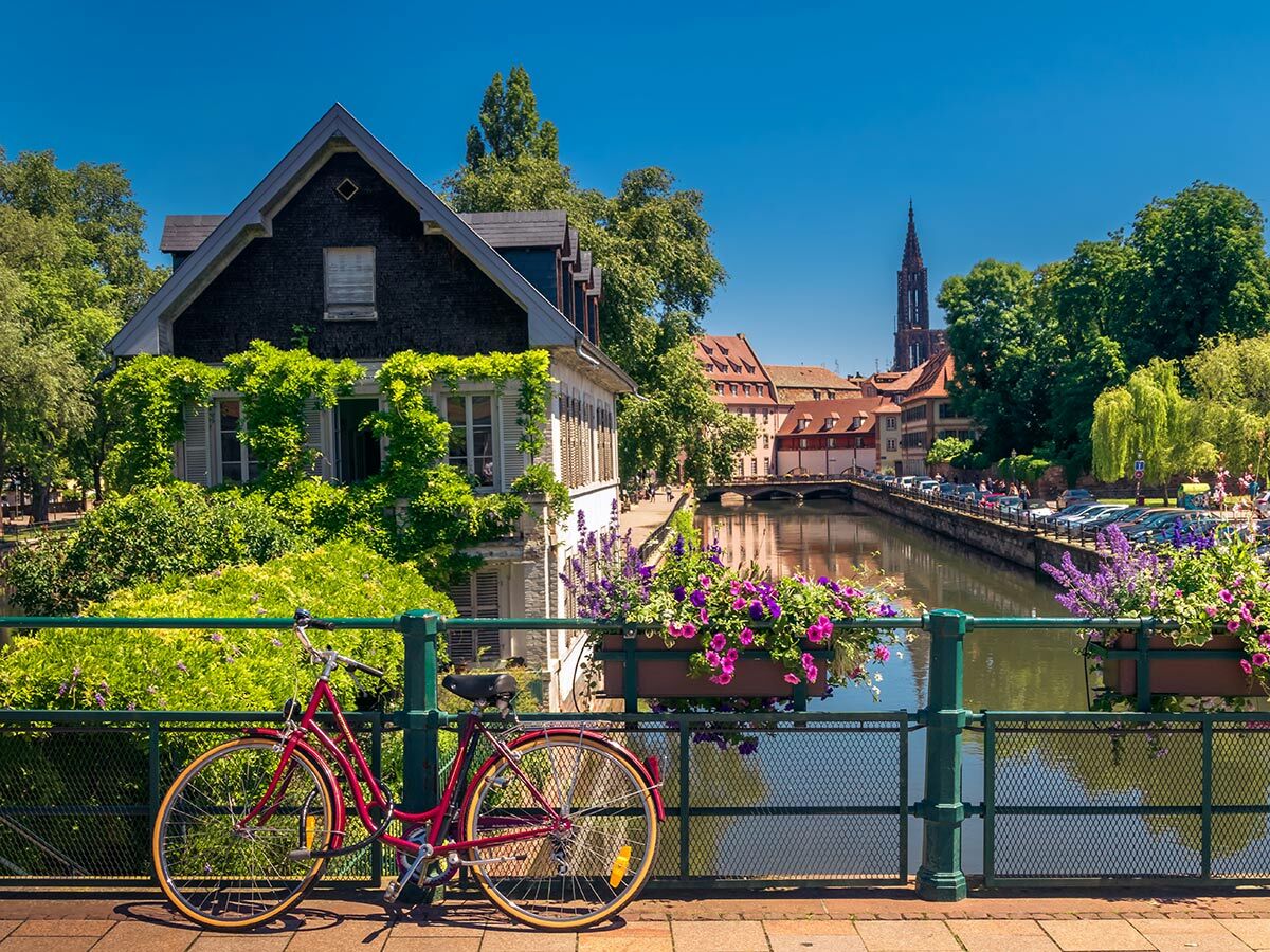 Rotes Fahrrad an Brücke mit Blumenkästen, Blick auf Kanal, grüne Bäume, historische Häuser und Straßburger Münster.