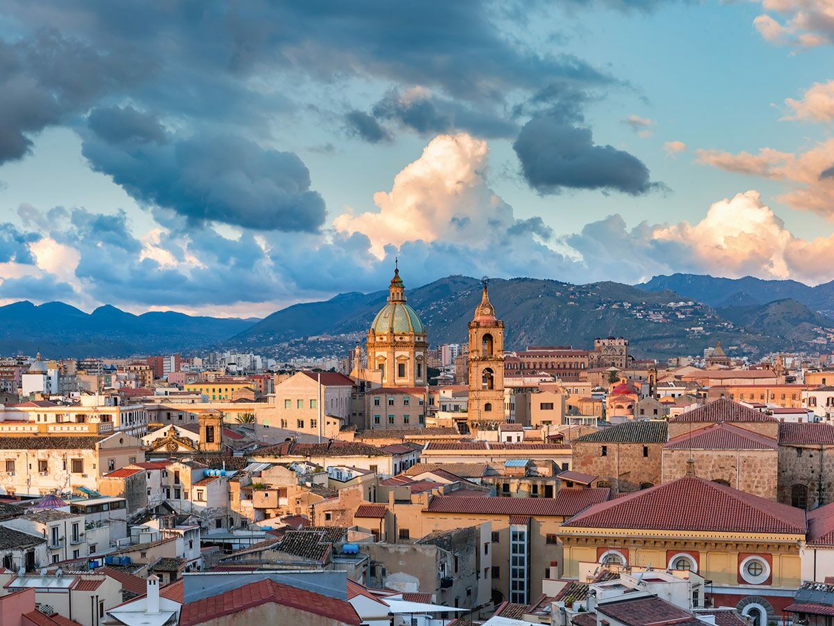 Palermo Skyline mit Kathedralenkuppel, historischen Gebäuden und Bergen im Hintergrund bei goldenem Sonnenuntergangslicht und dramatischen Wolken.