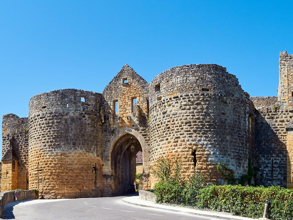 Ein imposantes mittelalterliches Burgtor aus Stein mit zwei runden Türmen und einem Torbogen, darunter eine Straße, vor blauem Himmel.