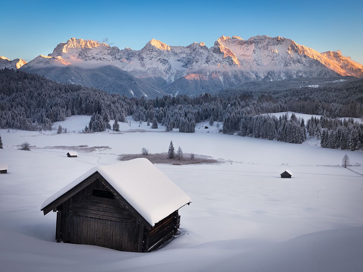 Atemberaubende Winterlandschaft mit schneebedeckten Bergen, Wald, gefrorenem See und Holzhütten bei Sonnenaufgang.